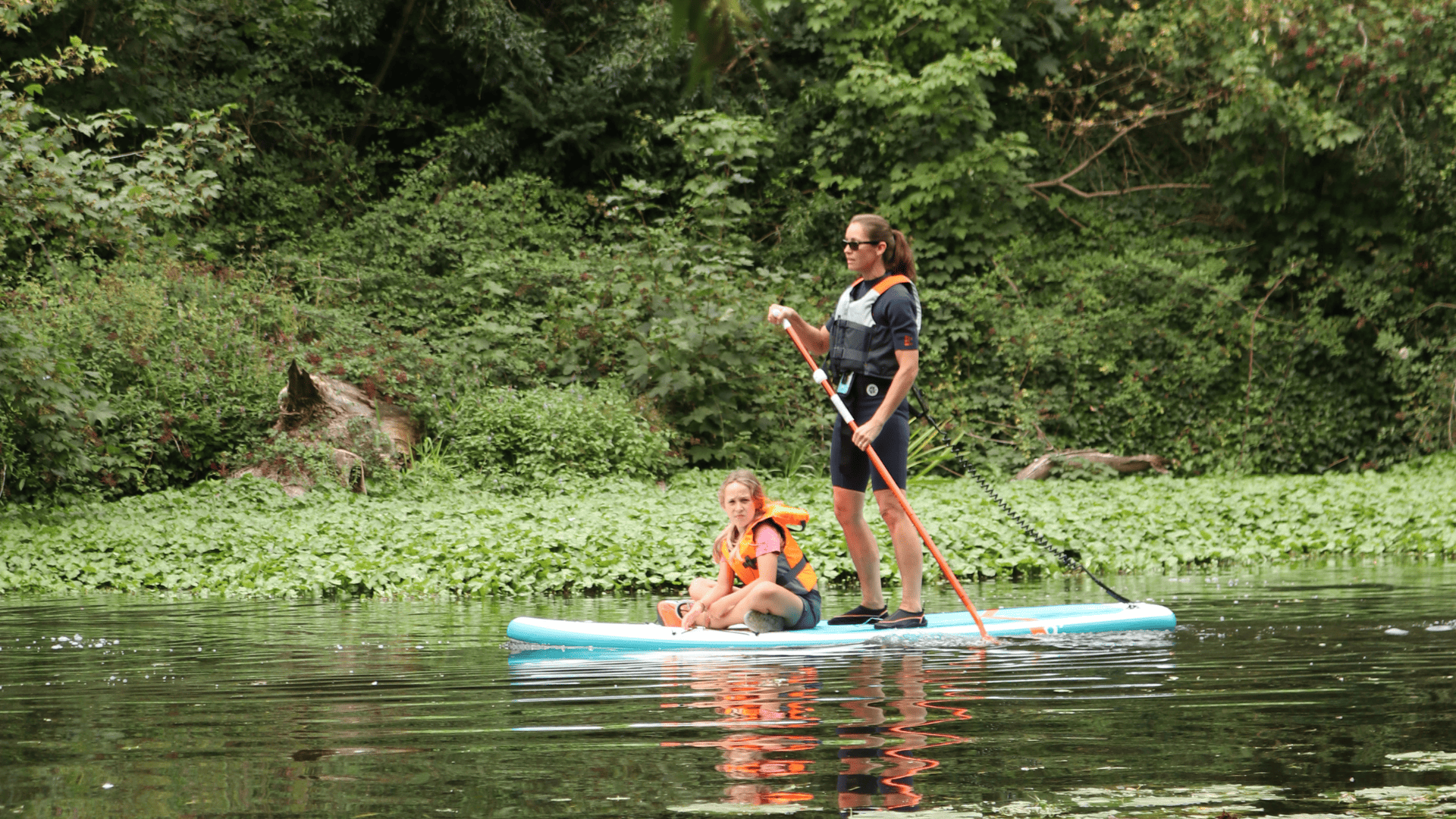 Woman paddleboarding with a young girl on the front of the board among green spaces. Digital membership.