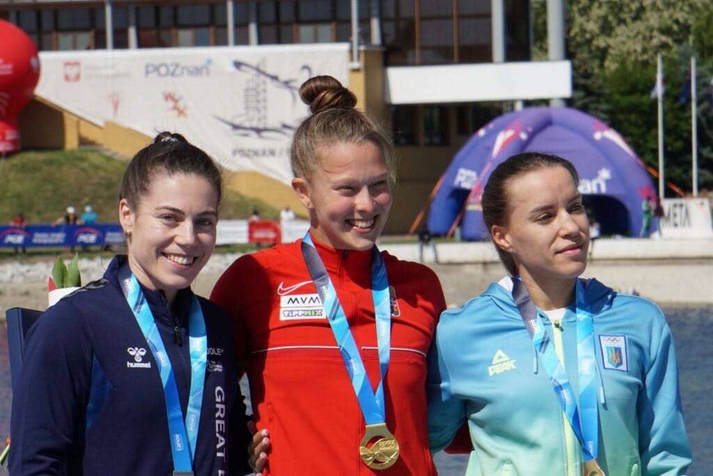 Beth Gill stands on the left, she is wearing a medal with a light blue ribbon around her nicek. She is white with dark brown hair tied in a high pony tail. She is wearing a dark blue zip up top and is with two other athletes on the podium 