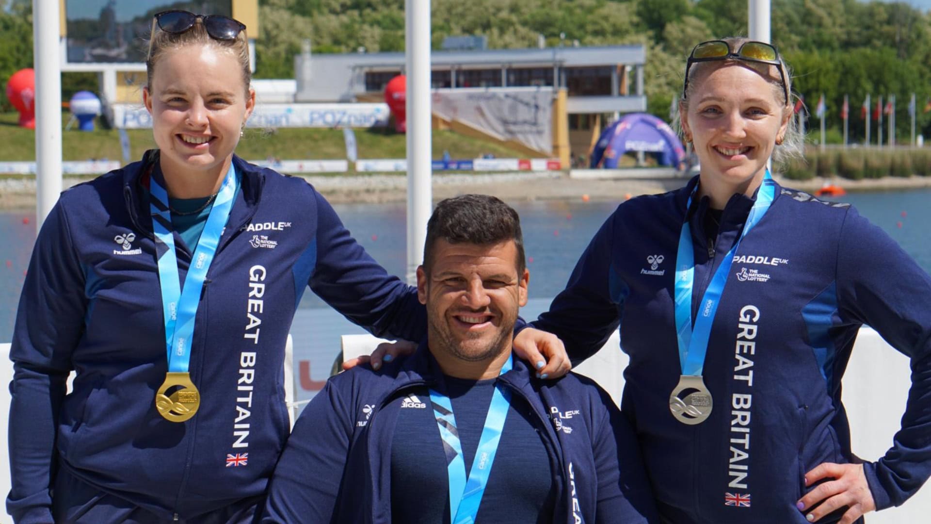 Hope Gordon stands on the left with her right arm resting on the right shoulder of Dave Phillipson who is sitting. Charlotte Henshaw has her left arm behind Dave's back. Hope is white with blonde ahir tied back and sunglasses on the top of her head, she is wearing a gold medal with a light blue ribbon around her neck. Charlotte is white with blonde hair tied back with sunglasses on the top of her head, she is wearing a silver medal with a light blue ribbon around her neck. Dave is white with short dark brown hair with a small fringe, he is wearing a light blue ribbon around his neck. All three are wearing dark blue quarter-zip tops with Great Britain written up the side of the body in white writing. They are standing in front of a marina