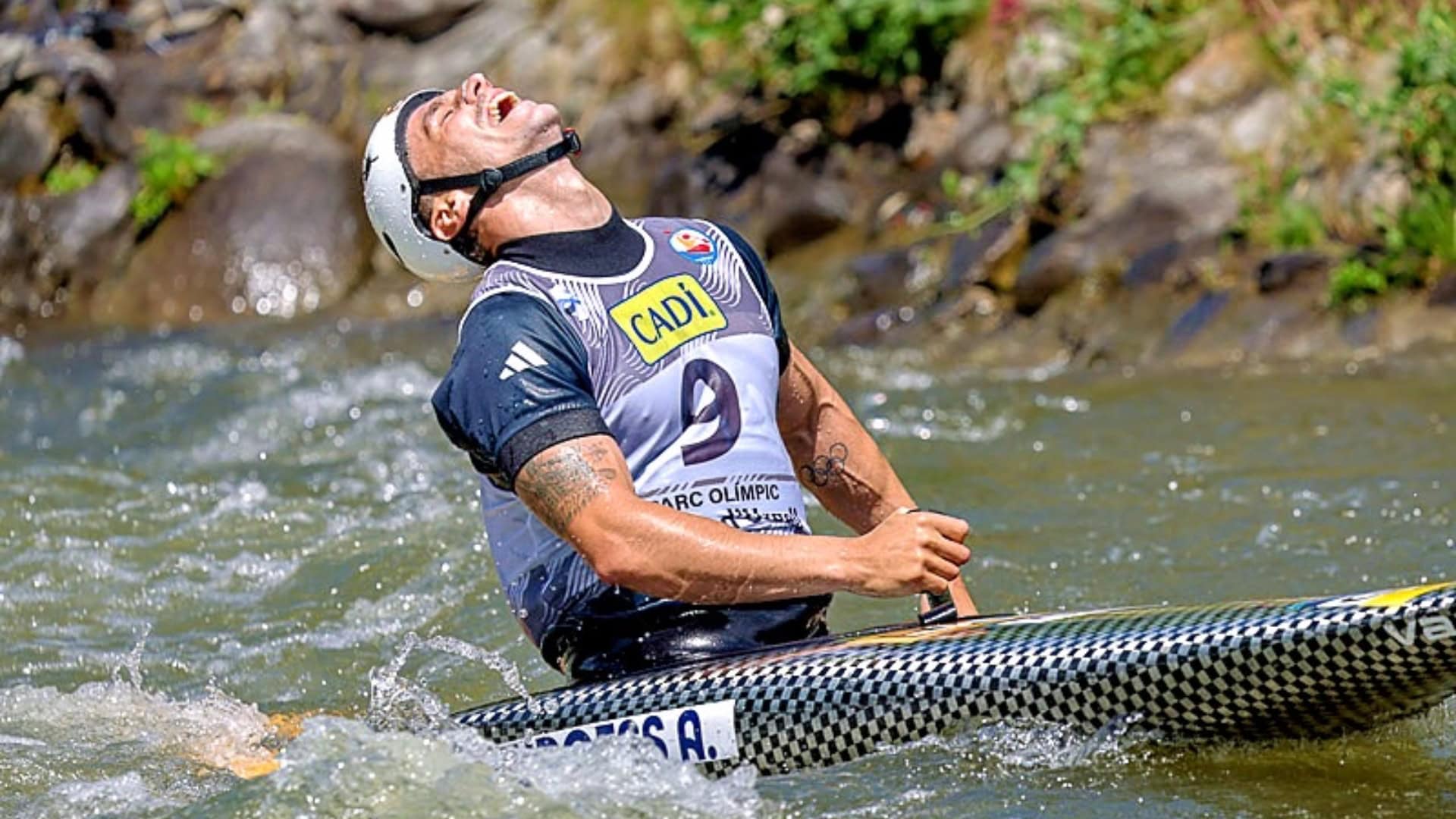 Adam Burgess looks up to the sky with an open mouth. He is sitting in a canoe on a white water course. He is wearing a white helmet with a black chin strap, he wears a navy blue short sleeved adidas top and a white vest with the number nine on it. His canoe is black with a geometric design.