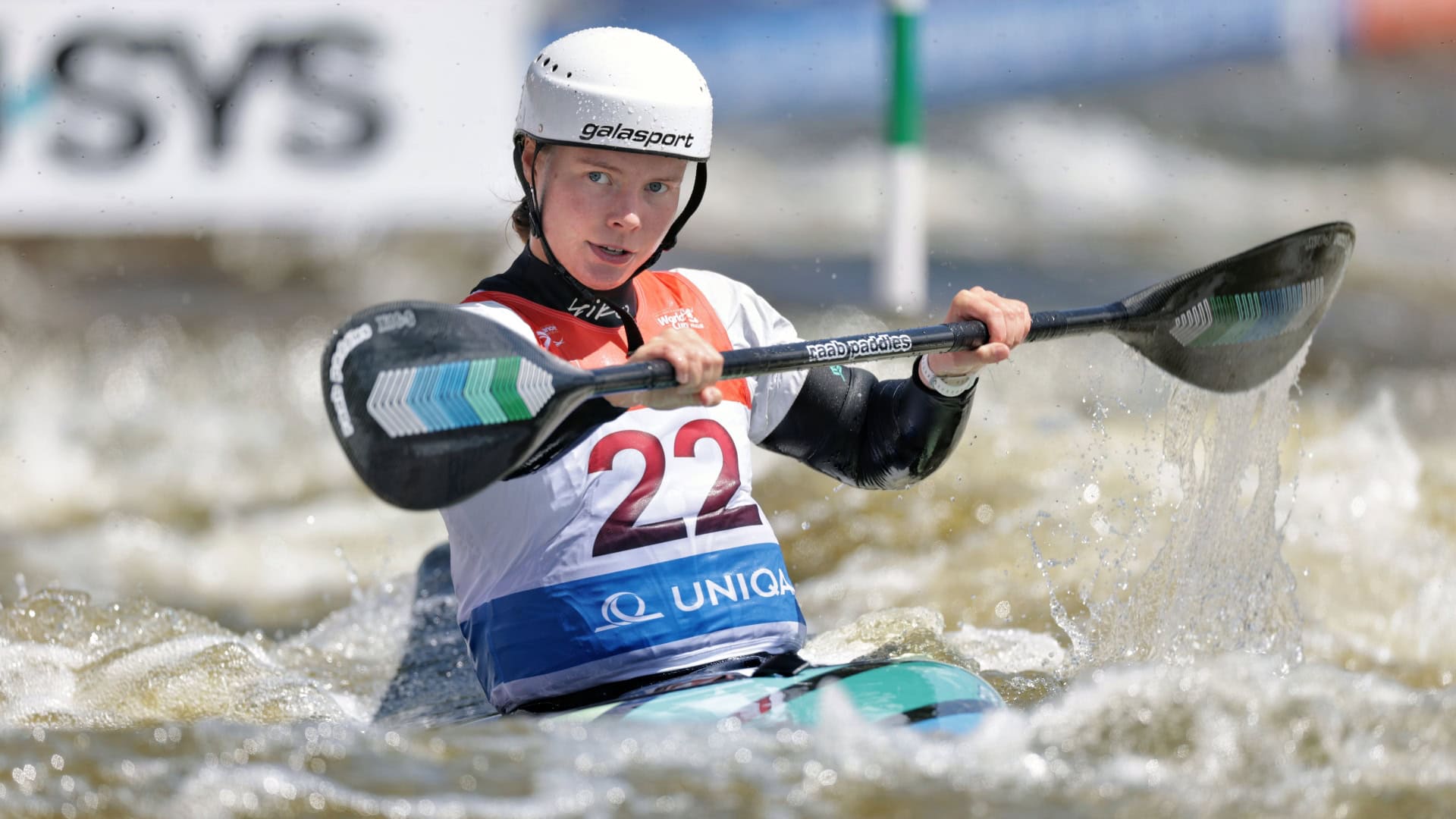 Lois Leaver competes in the women's Kayak cross in Prague. She is white with her hair pulled back. She is wearing a white helmet with black chinstrap. She wears a white t-shirt, black long sleeve shirt and a red blue and white bib with the number 22 on. She is holding a black kayak paddle and is in a multicoloured kayak on the water.