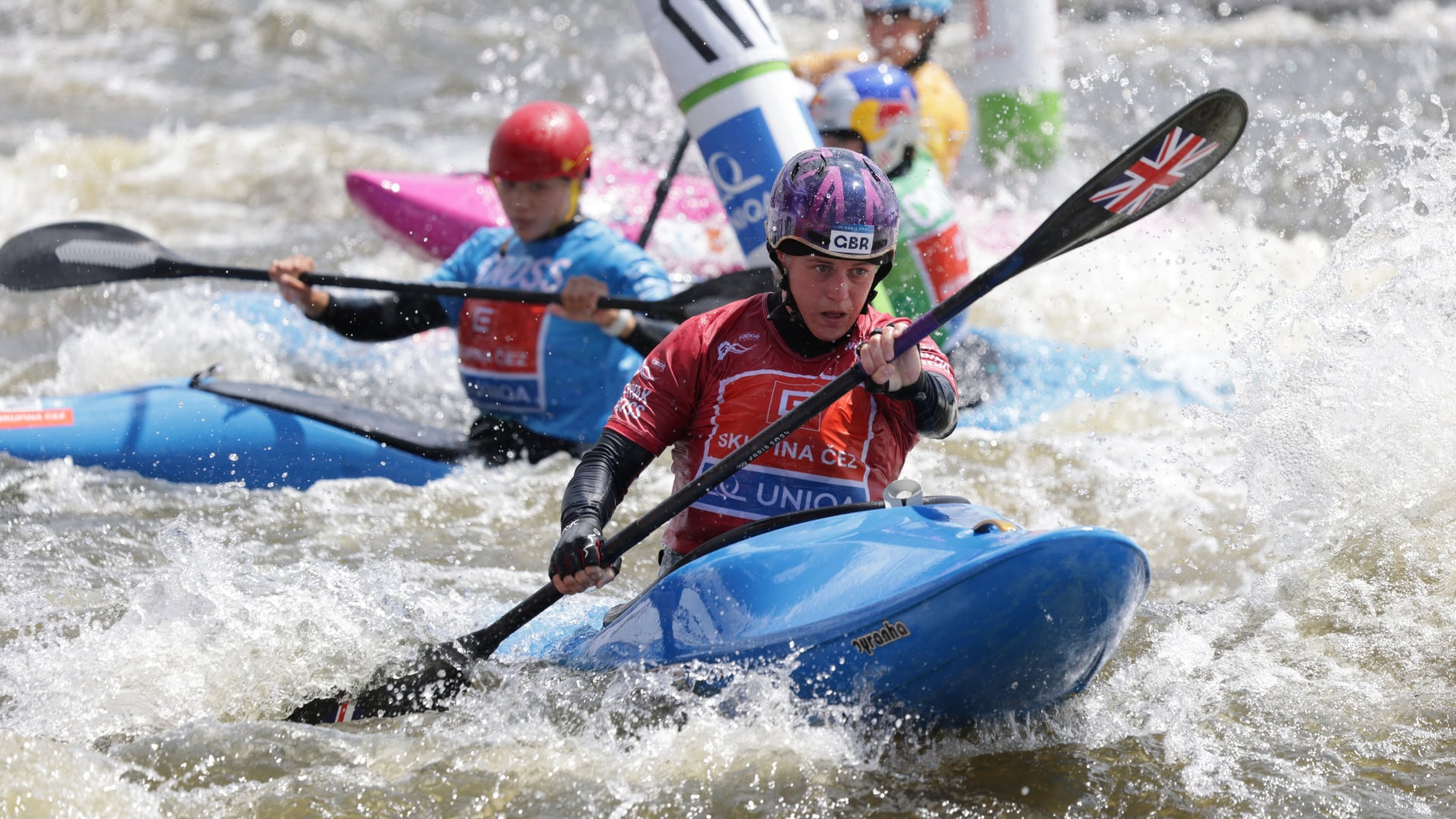 Kimberley Woods competes in the women's kayak cross at the Prague World Cup. She wears a pruple patterned helmet with black straps, a red short sleeve shirt and black long sleeve shirt under it. She is in a blue kayak and is holding a blue kayak paddle.