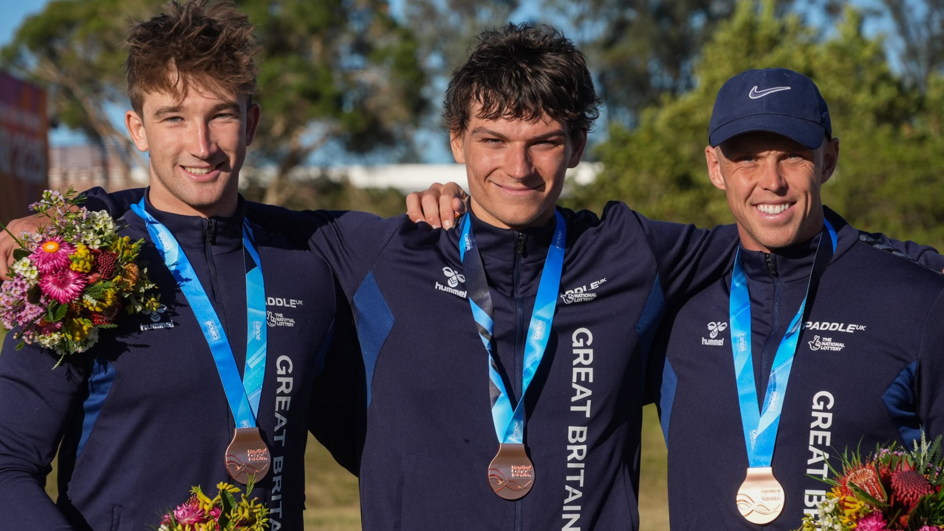 Ben Haylett, Jonny Dickson, Joe Clarke, stand with their arms around each other. Ben and Jonny have short brown hair, Joe is wearing a blue cap. All three are wearing navy zip jackets and bronze medals. They are each holding a bunch of flowers in their spare hand.